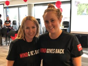 Two Wind River team members smile, wearing a t shirt that reads: Wind Gives Back written in white and red 