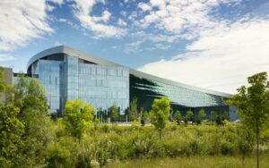 An image of a building that has a modern, curved architectural design. The windows and glass of the building reflects a partially cloudy day. 
