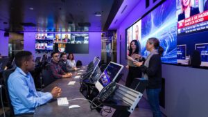 Two presenters are presenting to a small group seated at individual computer monitors. 