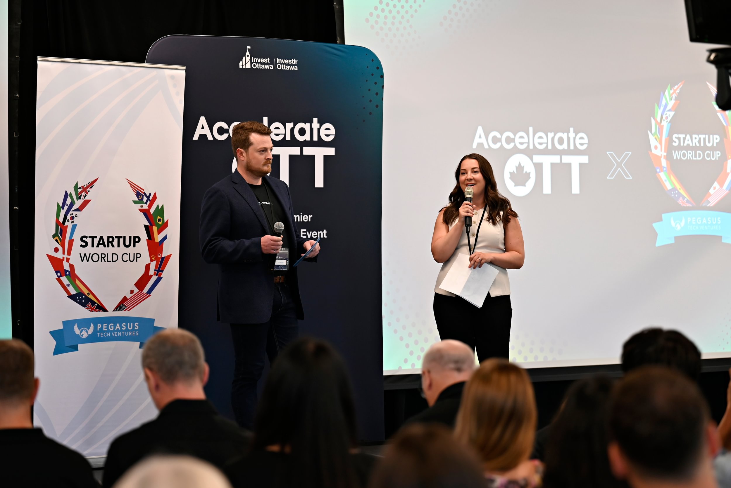 Two presenters speak on stage at an AccelerateOTT Startup World Cup event, addressing an audience during a pitch competition.