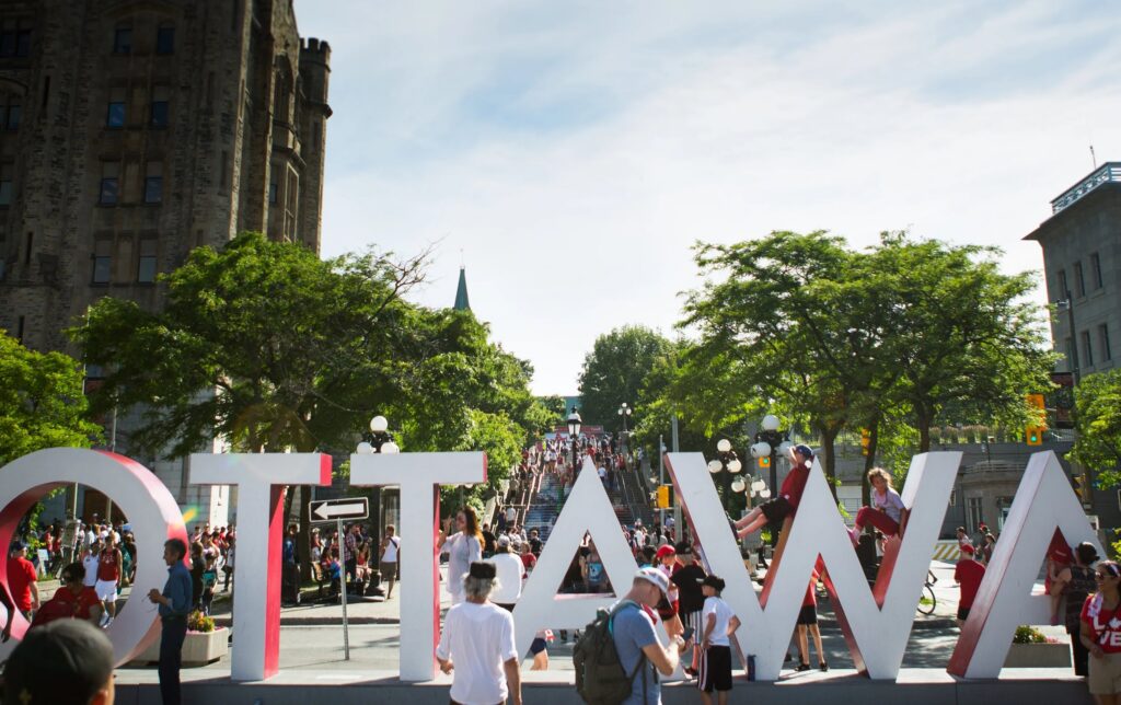 Crowds gather around the large “OTTAWA” sign on a sunny day, with historic buildings, trees, and a busy staircase in the background.