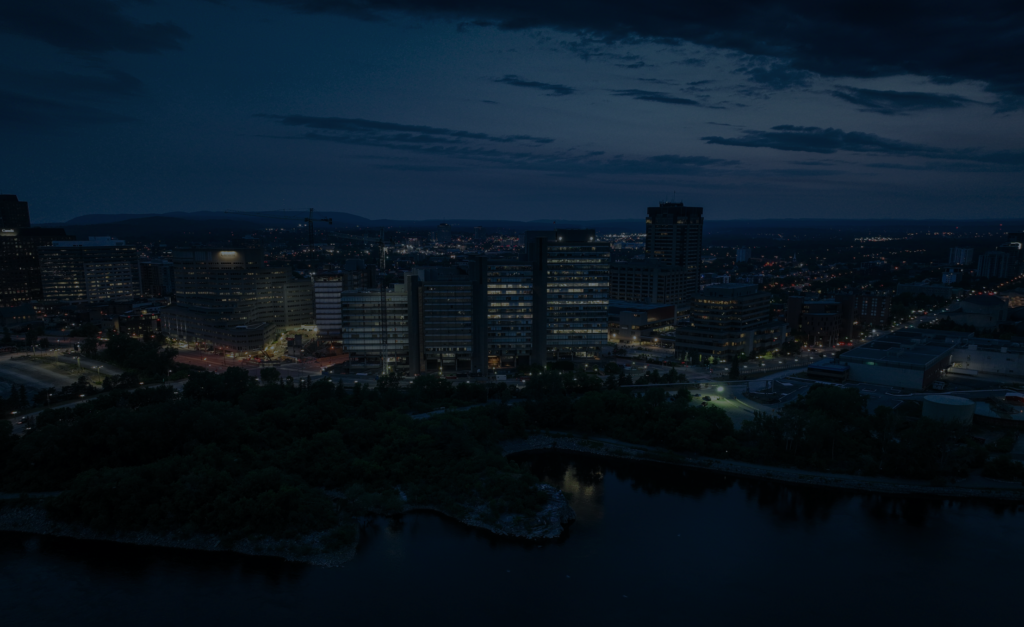 Aerial view of Ottawa’s downtown skyline at dusk, with illuminated office buildings and streets along the river, showcasing the city’s modern infrastructure and urban landscape.
